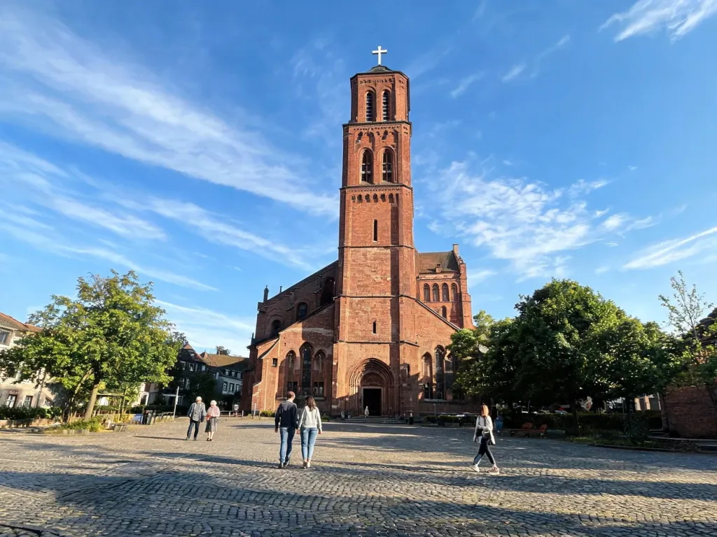 Wierni w słoneczne popołudnie przed zabytkowym kościołem Podwyższenia Krzyża Świętego w Poznaniu, spokojna atmosfera.